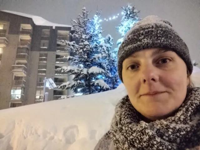 Orna Ní Choileáin faoi chaipín olla agus é ag cur sneachta go trom, crainn faoi shneachta taobh thiar di.